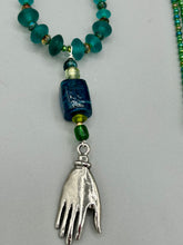 Load image into Gallery viewer, Bring the beauty of nature to your wardrobe with this gorgeous blue-green glass beaded necklace. Featuring a large rectangular blue-green glass bead, a lucky hand charm, mixed flat matte teal glass beads, and a hook-eye clasp for secure wear, this nature-inspired accessory adds a touch of summer to any outfit. Perfect for day-to-night looks.   Measurements:  Necklace: 19", Lucky hand pendant 3 1/2"