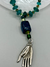 Load image into Gallery viewer, Bring the beauty of nature to your wardrobe with this gorgeous blue-green glass beaded necklace. Featuring a large rectangular blue-green glass bead, a lucky hand charm, mixed flat matte teal glass beads, and a hook-eye clasp for secure wear, this nature-inspired accessory adds a touch of summer to any outfit. Perfect for day-to-night looks.   Measurements:  Necklace: 19", Lucky hand pendant 3 1/2"