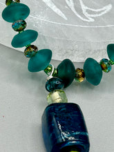 Load image into Gallery viewer, Bring the beauty of nature to your wardrobe with this gorgeous blue-green glass beaded necklace. Featuring a large rectangular blue-green glass bead, a lucky hand charm, mixed flat matte teal glass beads, and a hook-eye clasp for secure wear, this nature-inspired accessory adds a touch of summer to any outfit. Perfect for day-to-night looks.   Measurements:  Necklace: 19", Lucky hand pendant 3 1/2"