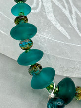 Load image into Gallery viewer, Bring the beauty of nature to your wardrobe with this gorgeous blue-green glass beaded necklace. Featuring a large rectangular blue-green glass bead, a lucky hand charm, mixed flat matte teal glass beads, and a hook-eye clasp for secure wear, this nature-inspired accessory adds a touch of summer to any outfit. Perfect for day-to-night looks.   Measurements:  Necklace: 19", Lucky hand pendant 3 1/2"
