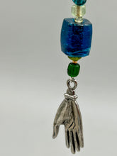 Load image into Gallery viewer, Bring the beauty of nature to your wardrobe with this gorgeous blue-green glass beaded necklace. Featuring a large rectangular blue-green glass bead, a lucky hand charm, mixed flat matte teal glass beads, and a hook-eye clasp for secure wear, this nature-inspired accessory adds a touch of summer to any outfit. Perfect for day-to-night looks.   Measurements:  Necklace: 19", Lucky hand pendant 3 1/2"
