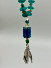 Load image into Gallery viewer, Bring the beauty of nature to your wardrobe with this gorgeous blue-green glass beaded necklace. Featuring a large rectangular blue-green glass bead, a lucky hand charm, mixed flat matte teal glass beads, and a hook-eye clasp for secure wear, this nature-inspired accessory adds a touch of summer to any outfit. Perfect for day-to-night looks.   Measurements:  Necklace: 19", Lucky hand pendant 3 1/2"