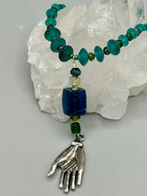 Load image into Gallery viewer, Bring the beauty of nature to your wardrobe with this gorgeous blue-green glass beaded necklace. Featuring a large rectangular blue-green glass bead, a lucky hand charm, mixed flat matte teal glass beads, and a hook-eye clasp for secure wear, this nature-inspired accessory adds a touch of summer to any outfit. Perfect for day-to-night looks.   Measurements:  Necklace: 19", Lucky hand pendant 3 1/2"
