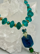 Load image into Gallery viewer, Bring the beauty of nature to your wardrobe with this gorgeous blue-green glass beaded necklace. Featuring a large rectangular blue-green glass bead, a lucky hand charm, mixed flat matte teal glass beads, and a hook-eye clasp for secure wear, this nature-inspired accessory adds a touch of summer to any outfit. Perfect for day-to-night looks.   Measurements:  Necklace: 19", Lucky hand pendant 3 1/2"