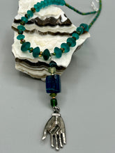 Load image into Gallery viewer, Bring the beauty of nature to your wardrobe with this gorgeous blue-green glass beaded necklace. Featuring a large rectangular blue-green glass bead, a lucky hand charm, mixed flat matte teal glass beads, and a hook-eye clasp for secure wear, this nature-inspired accessory adds a touch of summer to any outfit. Perfect for day-to-night looks.   Measurements:  Necklace: 19", Lucky hand pendant 3 1/2"