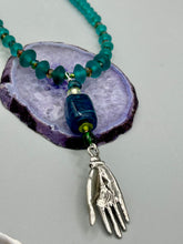 Load image into Gallery viewer, Bring the beauty of nature to your wardrobe with this gorgeous blue-green glass beaded necklace. Featuring a large rectangular blue-green glass bead, a lucky hand charm, mixed flat matte teal glass beads, and a hook-eye clasp for secure wear, this nature-inspired accessory adds a touch of summer to any outfit. Perfect for day-to-night looks.   Measurements:  Necklace: 19", Lucky hand pendant 3 1/2"