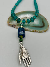 Load image into Gallery viewer, Bring the beauty of nature to your wardrobe with this gorgeous blue-green glass beaded necklace. Featuring a large rectangular blue-green glass bead, a lucky hand charm, mixed flat matte teal glass beads, and a hook-eye clasp for secure wear, this nature-inspired accessory adds a touch of summer to any outfit. Perfect for day-to-night looks.   Measurements:  Necklace: 19", Lucky hand pendant 3 1/2"