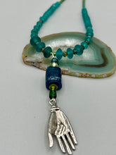 Load image into Gallery viewer, Bring the beauty of nature to your wardrobe with this gorgeous blue-green glass beaded necklace. Featuring a large rectangular blue-green glass bead, a lucky hand charm, mixed flat matte teal glass beads, and a hook-eye clasp for secure wear, this nature-inspired accessory adds a touch of summer to any outfit. Perfect for day-to-night looks.   Measurements:  Necklace: 19", Lucky hand pendant 3 1/2"