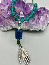 Load image into Gallery viewer, Bring the beauty of nature to your wardrobe with this gorgeous blue-green glass beaded necklace. Featuring a large rectangular blue-green glass bead, a lucky hand charm, mixed flat matte teal glass beads, and a hook-eye clasp for secure wear, this nature-inspired accessory adds a touch of summer to any outfit. Perfect for day-to-night looks.   Measurements:  Necklace: 19", Lucky hand pendant 3 1/2"