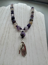 Load image into Gallery viewer, This nature-inspired accessory features an array of unique hues, including rainbow fluorite stone beads and iridescent dark shimmery purple glass beads with hints of gold, blue, pink, and green. The secure hook-eye clasp makes it perfect for day to night wear, allowing you to add a touch of summer to your wardrobe. The necklace is further complemented by a large square-shaped rainbow fluorite bead and a lucky hand charm. Measurements: Necklace: 18", Lucky hand pendant 3"
