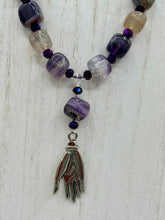Load image into Gallery viewer, This nature-inspired accessory features an array of unique hues, including rainbow fluorite stone beads and iridescent dark shimmery purple glass beads with hints of gold, blue, pink, and green. The secure hook-eye clasp makes it perfect for day to night wear, allowing you to add a touch of summer to your wardrobe. The necklace is further complemented by a large square-shaped rainbow fluorite bead and a lucky hand charm. Measurements: Necklace: 18", Lucky hand pendant 3"