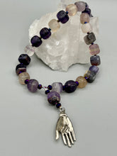 Load image into Gallery viewer, This nature-inspired accessory features an array of unique hues, including rainbow fluorite stone beads and iridescent dark shimmery purple glass beads with hints of gold, blue, pink, and green. The secure hook-eye clasp makes it perfect for day to night wear, allowing you to add a touch of summer to your wardrobe. The necklace is further complemented by a large square-shaped rainbow fluorite bead and a lucky hand charm. Measurements: Necklace: 18", Lucky hand pendant 3"