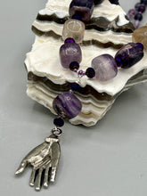Load image into Gallery viewer, This nature-inspired accessory features an array of unique hues, including rainbow fluorite stone beads and iridescent dark shimmery purple glass beads with hints of gold, blue, pink, and green. The secure hook-eye clasp makes it perfect for day to night wear, allowing you to add a touch of summer to your wardrobe. The necklace is further complemented by a large square-shaped rainbow fluorite bead and a lucky hand charm. Measurements: Necklace: 18", Lucky hand pendant 3"