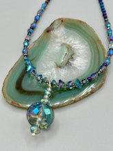 Load image into Gallery viewer, Make a bold statement with this shimmery aqua blue and blue tones glass beaded necklace. The round faceted shaped pendant compliments the cool tones of the necklace, perfect for adding a rich pop of color to any Spring outfit. The draped design gives this piece an elegant, bohemian look. Measurements: Necklace is 18” in length, pendant is 1 1/2”