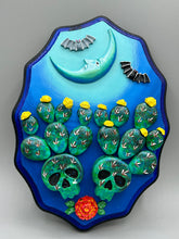 Load image into Gallery viewer, This carefully crafted polymer clay wall art showcases a wood frame adorned with a crescent moon motif. A striking color palette of deep sky tones, including light blue and royal blue to black, further enhances the piece. Delicate cacti plants bloom with light yellow flowers, with spooky skulls are incorporated among the cacti plant and a finishing touch of flying bat motifs. Measurements: H 9" x W 6 1/2” x D 1 1/4"