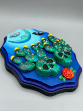 Load image into Gallery viewer, This carefully crafted polymer clay wall art showcases a wood frame adorned with a crescent moon motif. A striking color palette of deep sky tones, including light blue and royal blue to black, further enhances the piece. Delicate cacti plants bloom with light yellow flowers, with spooky skulls are incorporated among the cacti plant and a finishing touch of flying bat motifs. Measurements: H 9" x W 6 1/2” x D 1 1/4"