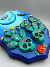 Load image into Gallery viewer, This carefully crafted polymer clay wall art showcases a wood frame adorned with a crescent moon motif. A striking color palette of deep sky tones, including light blue and royal blue to black, further enhances the piece. Delicate cacti plants bloom with light yellow flowers, with spooky skulls are incorporated among the cacti plant and a finishing touch of flying bat motifs. Measurements: H 9" x W 6 1/2” x D 1 1/4"