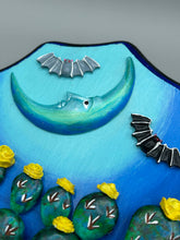 Load image into Gallery viewer, This carefully crafted polymer clay wall art showcases a wood frame adorned with a crescent moon motif. A striking color palette of deep sky tones, including light blue and royal blue to black, further enhances the piece. Delicate cacti plants bloom with light yellow flowers, with spooky skulls are incorporated among the cacti plant and a finishing touch of flying bat motifs. Measurements: H 9" x W 6 1/2” x D 1 1/4"