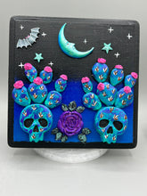 Load image into Gallery viewer, This carefully crafted polymer clay wall art showcases a wood frame adorned with a crescent moon and stars motifs. A striking dark palette of deep sky tones, including royal blue to black, further enhances the piece. Delicate cacti plants bloom with light pink/purple flowers, with spooky skulls are incorporated among the cacti plant and a finishing touch of a tiny flying bat motif. Measurements: H 6 3/4" x W 6 3/4” x D 1 1/4"