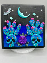 Load image into Gallery viewer, This carefully crafted polymer clay wall art showcases a wood frame adorned with a crescent moon and stars motifs. A striking dark palette of deep sky tones, including royal blue to black, further enhances the piece. Delicate cacti plants bloom with light pink/purple flowers, with spooky skulls are incorporated among the cacti plant and a finishing touch of a tiny flying bat motif. Measurements: H 6 3/4" x W 6 3/4” x D 1 1/4"