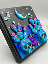 Load image into Gallery viewer, This carefully crafted polymer clay wall art showcases a wood frame adorned with a crescent moon and stars motifs. A striking dark palette of deep sky tones, including royal blue to black, further enhances the piece. Delicate cacti plants bloom with light pink/purple flowers, with spooky skulls are incorporated among the cacti plant and a finishing touch of a tiny flying bat motif. Measurements: H 6 3/4" x W 6 3/4” x D 1 1/4"