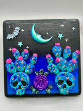 Load image into Gallery viewer, This carefully crafted polymer clay wall art showcases a wood frame adorned with a crescent moon and stars motifs. A striking dark palette of deep sky tones, including royal blue to black, further enhances the piece. Delicate cacti plants bloom with light pink/purple flowers, with spooky skulls are incorporated among the cacti plant and a finishing touch of a tiny flying bat motif. Measurements: H 6 3/4" x W 6 3/4” x D 1 1/4"