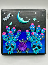 Load image into Gallery viewer, This carefully crafted polymer clay wall art showcases a wood frame adorned with a crescent moon and stars motifs. A striking dark palette of deep sky tones, including royal blue to black, further enhances the piece. Delicate cacti plants bloom with light pink/purple flowers, with spooky skulls are incorporated among the cacti plant and a finishing touch of a tiny flying bat motif. Measurements: H 6 3/4" x W 6 3/4” x D 1 1/4"