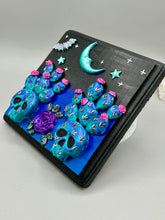 Load image into Gallery viewer, This carefully crafted polymer clay wall art showcases a wood frame adorned with a crescent moon and stars motifs. A striking dark palette of deep sky tones, including royal blue to black, further enhances the piece. Delicate cacti plants bloom with light pink/purple flowers, with spooky skulls are incorporated among the cacti plant and a finishing touch of a tiny flying bat motif. Measurements: H 6 3/4" x W 6 3/4” x D 1 1/4"
