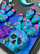 Load image into Gallery viewer, This carefully crafted polymer clay wall art showcases a wood frame adorned with a crescent moon and stars motifs. A striking dark palette of deep sky tones, including royal blue to black, further enhances the piece. Delicate cacti plants bloom with light pink/purple flowers, with spooky skulls are incorporated among the cacti plant and a finishing touch of a tiny flying bat motif. Measurements: H 6 3/4" x W 6 3/4” x D 1 1/4"