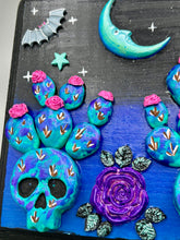 Load image into Gallery viewer, This carefully crafted polymer clay wall art showcases a wood frame adorned with a crescent moon and stars motifs. A striking dark palette of deep sky tones, including royal blue to black, further enhances the piece. Delicate cacti plants bloom with light pink/purple flowers, with spooky skulls are incorporated among the cacti plant and a finishing touch of a tiny flying bat motif. Measurements: H 6 3/4" x W 6 3/4” x D 1 1/4"