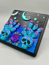 Load image into Gallery viewer, This carefully crafted polymer clay wall art showcases a wood frame adorned with a crescent moon and stars motifs. A striking dark palette of deep sky tones, including royal blue to black, further enhances the piece. Delicate cacti plants bloom with light pink/purple flowers, with spooky skulls are incorporated among the cacti plant and a finishing touch of a tiny flying bat motif. Measurements: H 6 3/4" x W 6 3/4” x D 1 1/4"