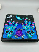 Load image into Gallery viewer, This carefully crafted polymer clay wall art showcases a wood frame adorned with a crescent moon and stars motifs. A striking dark palette of deep sky tones, including royal blue to black, further enhances the piece. Delicate cacti plants bloom with light pink/purple flowers, with spooky skulls are incorporated among the cacti plant and a finishing touch of a tiny flying bat motif. Measurements: H 6 3/4" x W 6 3/4” x D 1 1/4"