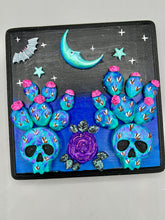 Load image into Gallery viewer, This carefully crafted polymer clay wall art showcases a wood frame adorned with a crescent moon and stars motifs. A striking dark palette of deep sky tones, including royal blue to black, further enhances the piece. Delicate cacti plants bloom with light pink/purple flowers, with spooky skulls are incorporated among the cacti plant and a finishing touch of a tiny flying bat motif. Measurements: H 6 3/4" x W 6 3/4” x D 1 1/4"