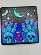 Load image into Gallery viewer, This carefully crafted polymer clay wall art showcases a wood frame adorned with a crescent moon and stars motifs. A striking dark palette of deep sky tones, including royal blue to black, further enhances the piece. Delicate cacti plants bloom with light pink/purple flowers, with spooky skulls are incorporated among the cacti plant and a finishing touch of a tiny flying bat motif. Measurements: H 6 3/4" x W 6 3/4” x D 1 1/4"
