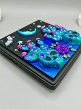 Load image into Gallery viewer, This carefully crafted polymer clay wall art showcases a wood frame adorned with a crescent moon and stars motifs. A striking dark palette of deep sky tones, including royal blue to black, further enhances the piece. Delicate cacti plants bloom with light pink/purple flowers, with spooky skulls are incorporated among the cacti plant and a finishing touch of a tiny flying bat motif. Measurements: H 6 3/4" x W 6 3/4” x D 1 1/4"