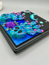 Load image into Gallery viewer, This carefully crafted polymer clay wall art showcases a wood frame adorned with a crescent moon and stars motifs. A striking dark palette of deep sky tones, including royal blue to black, further enhances the piece. Delicate cacti plants bloom with light pink/purple flowers, with spooky skulls are incorporated among the cacti plant and a finishing touch of a tiny flying bat motif. Measurements: H 6 3/4" x W 6 3/4” x D 1 1/4"