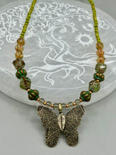 Load image into Gallery viewer, This intricately crafted pendant features a basil leaf molded in brass and adorned with stunningly warm greens, blues, and gold tone beaded necklace. Its eye-catching butterfly design adds a bold, yet whimsical touch, making it an ideal choice for a summer look. This beaded necklace is crafted from a combination of brass floral beads with a subtle patina hue, champagne melon beads, and dark, faceted avocado green pressed glass beads.  Measurements: Necklace is 18 1/2” in length - pendant is 1” Height