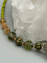 Load image into Gallery viewer, This intricately crafted pendant features a basil leaf molded in brass and adorned with stunningly warm greens, blues, and gold tone beaded necklace. Its eye-catching butterfly design adds a bold, yet whimsical touch, making it an ideal choice for a summer look. This beaded necklace is crafted from a combination of brass floral beads with a subtle patina hue, champagne melon beads, and dark, faceted avocado green pressed glass beads.  Measurements: Necklace is 18 1/2” in length - pendant is 1” Height