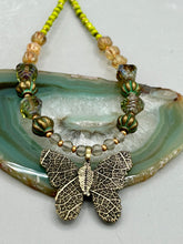 Load image into Gallery viewer, This intricately crafted pendant features a basil leaf molded in brass and adorned with stunningly warm greens, blues, and gold tone beaded necklace. Its eye-catching butterfly design adds a bold, yet whimsical touch, making it an ideal choice for a summer look. This beaded necklace is crafted from a combination of brass floral beads with a subtle patina hue, champagne melon beads, and dark, faceted avocado green pressed glass beads.  Measurements: Necklace is 18 1/2” in length - pendant is 1” Height