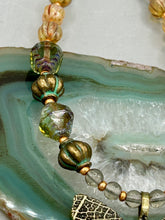 Load image into Gallery viewer, This intricately crafted pendant features a basil leaf molded in brass and adorned with stunningly warm greens, blues, and gold tone beaded necklace. Its eye-catching butterfly design adds a bold, yet whimsical touch, making it an ideal choice for a summer look. This beaded necklace is crafted from a combination of brass floral beads with a subtle patina hue, champagne melon beads, and dark, faceted avocado green pressed glass beads.  Measurements: Necklace is 18 1/2” in length - pendant is 1” Height