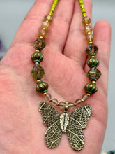 Load image into Gallery viewer, This intricately crafted pendant features a basil leaf molded in brass and adorned with stunningly warm greens, blues, and gold tone beaded necklace. Its eye-catching butterfly design adds a bold, yet whimsical touch, making it an ideal choice for a summer look. This beaded necklace is crafted from a combination of brass floral beads with a subtle patina hue, champagne melon beads, and dark, faceted avocado green pressed glass beads.  Measurements: Necklace is 18 1/2” in length - pendant is 1” Height