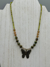 Load image into Gallery viewer, This intricately crafted pendant features a basil leaf molded in brass and adorned with stunningly warm greens, blues, and gold tone beaded necklace. Its eye-catching butterfly design adds a bold, yet whimsical touch, making it an ideal choice for a summer look. This beaded necklace is crafted from a combination of brass floral beads with a subtle patina hue, champagne melon beads, and dark, faceted avocado green pressed glass beads.  Measurements: Necklace is 18 1/2” in length - pendant is 1” Height