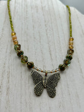 Load image into Gallery viewer, This intricately crafted pendant features a basil leaf molded in brass and adorned with stunningly warm greens, blues, and gold tone beaded necklace. Its eye-catching butterfly design adds a bold, yet whimsical touch, making it an ideal choice for a summer look. This beaded necklace is crafted from a combination of brass floral beads with a subtle patina hue, champagne melon beads, and dark, faceted avocado green pressed glass beads.  Measurements: Necklace is 18 1/2” in length - pendant is 1” Height