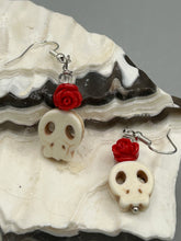Load image into Gallery viewer, These stylish skull earrings are perfect for adding a unique flair to any outfit. Whether you're looking for a Dia De Los Muertos accessory or simply looking to draw inspiration, these goth pieces perfectly capture the vampire goth, witchy, dark, Chingona Cabrona, Latina Chicana styles. Howlite stone skull bead with red quartzize floral beads.  Measurements: 1 1/4 ” in length