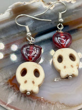 Load image into Gallery viewer, These stylish skull earrings are perfect for adding a unique flair to any outfit. Whether you're looking for a Dia De Los Muertos accessory or simply looking to draw inspiration, these goth pieces perfectly capture the vampire goth, witchy, dark, Chingona Cabrona, Latina Chicana styles. Howlite stone skull beaded earrings come with a ruby red heart-shaped glass beaded that has a very shimmery and vivid iridescent hue.  Measurements: 1 1/4 ” in length