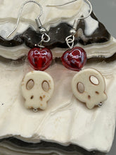Load image into Gallery viewer, These stylish skull earrings are perfect for adding a unique flair to any outfit. Whether you're looking for a Dia De Los Muertos accessory or simply looking to draw inspiration, these goth pieces perfectly capture the vampire goth, witchy, dark, Chingona Cabrona, Latina Chicana styles. Howlite stone skull beaded earrings come with a ruby red heart-shaped glass beaded that has a very shimmery and vivid iridescent hue.  Measurements: 1 1/4 ” in length