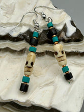 Load image into Gallery viewer, These stylish skull earrings are perfect for adding a unique flair to any outfit. Whether you're looking for a Dia De Los Muertos accessory or simply looking to draw inspiration, these goth pieces perfectly capture the vampire goth, witchy, dark, Chingona Cabrona, Latina Chicana styles. Skull-shaped bone beads adorned with dark brown, turquoise, and gold beaded earrings make up this unique set of earrings. Measurements: 1 1/2 ” in length