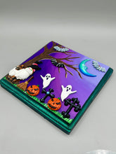 Load image into Gallery viewer, This captivating starry night calico kitty in the cemetery is artfully crafted with a black wood frame with a green iridescent finish and a dark purple hue background for an eye-catching and captivating nocturnal effect art piece. This kitty cat is dressed in a witch's hat while paying a visit to the cemetery accompanied by a broom and a jack-o-lantern and bat friends. This bewitching and whimsical Halloween-themed art piece adds a one-of-a-kind Halloween touch to any home or office.