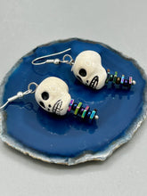 Load image into Gallery viewer, Peruvian off white ceramic skull beads come with rainbow hematite spine earrings. These stylish skull earrings are perfect for adding a unique flair to any outfit. Whether you're looking for a Dia De Los Muertos accessory or simply looking to draw inspiration, these goth pieces perfectly capture the vampire goth, witchy, dark, Chingona Cabrona, Latina Chicana styles. Measurements: 1 1/4” in length