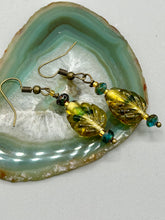 Load image into Gallery viewer, This pair of earrings features a dark amber and green tone, pressed glass leaf charm in an iridescent finish that gives off subtle hues of gold. Complemented with dark teal and gold glass beaded earrings, this set is sure to sparkle.  Measurements: 1 1/4" length